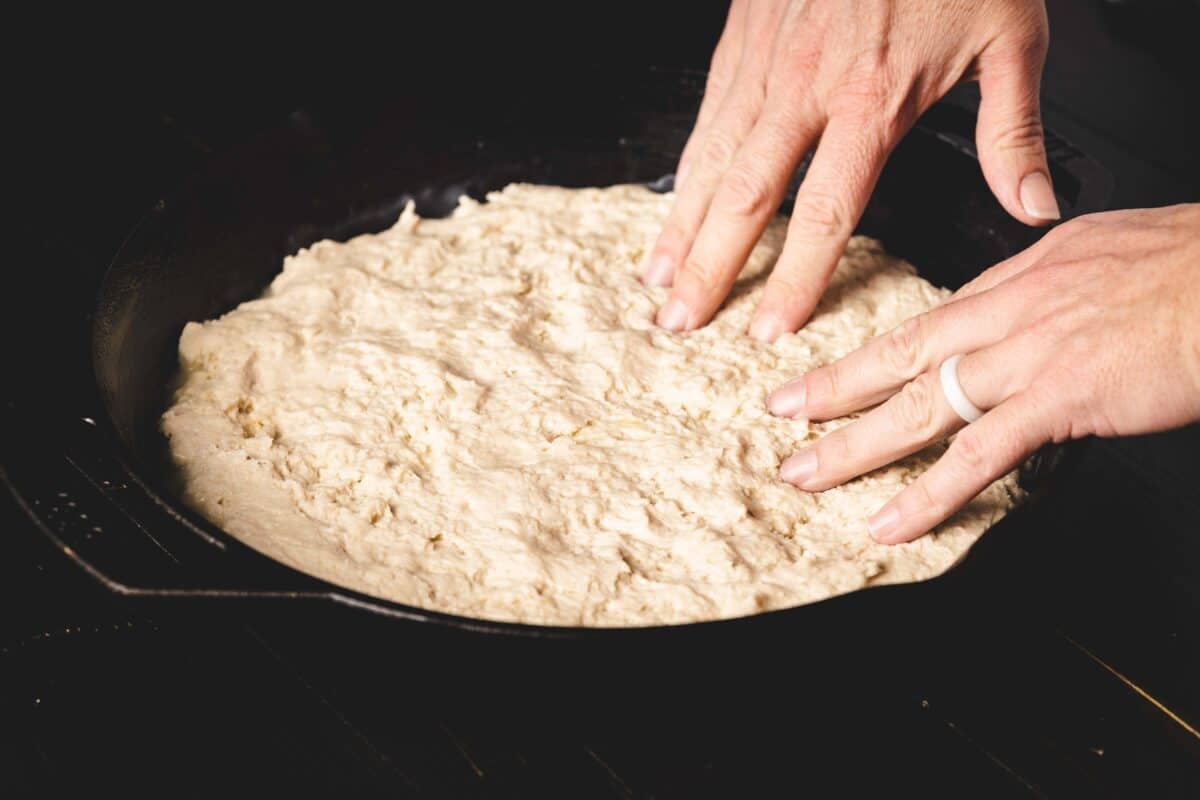 Two hands working dough into a cast iron skillet.