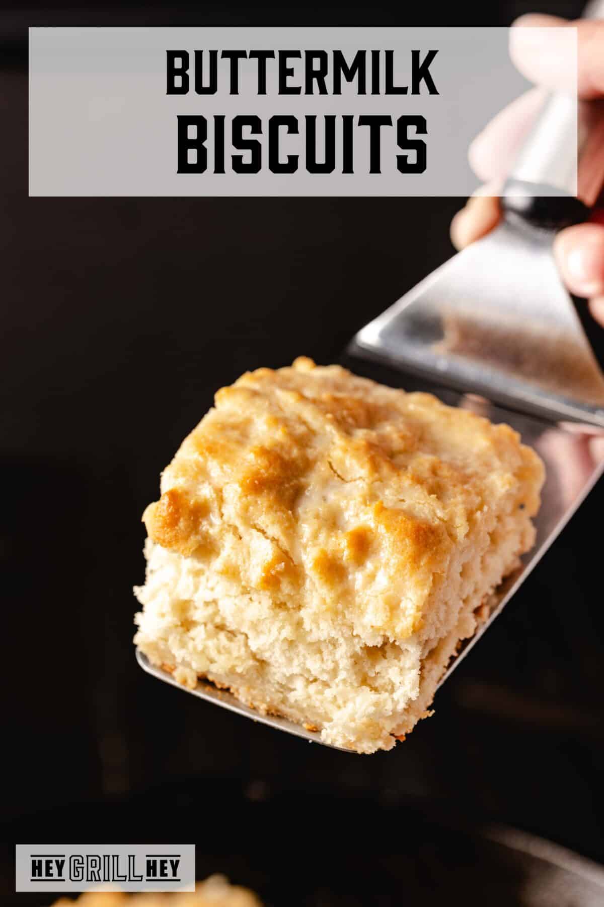 A metal spatula holding homemade biscuit over a black background. The text overlay reads "Buttermilk Biscuits" at the top, and "Hey Grill Hey" at the bottom.