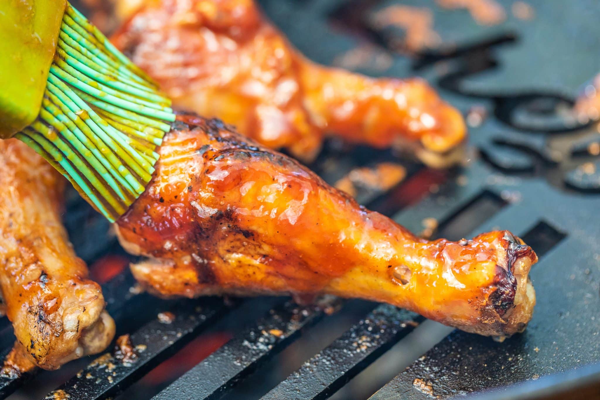 chicken drumsticks on a grill being basted with BBQ sauce by a basting brush.