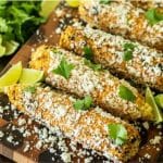 Mexican street corn on a wooden cutting board garnished with lime wedges and cilantro.