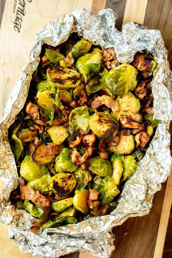 Overhead shot of Grilled Brussels and bacon in tinfoil. Displayed on a wood cutting board.