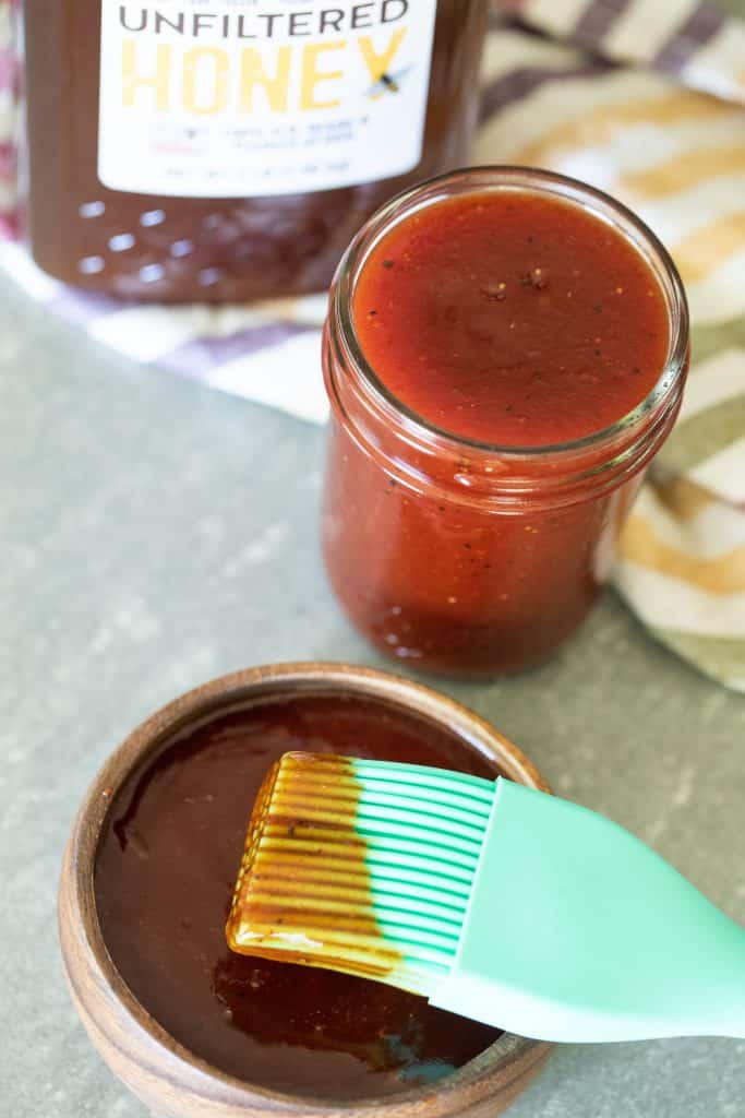 Mint colored basting brush dipped in honey barbecue sauce resting on a small wood bowl full of honey barbecue sauce with a small mason jar full of more sauce with a bottle of honey in the background.