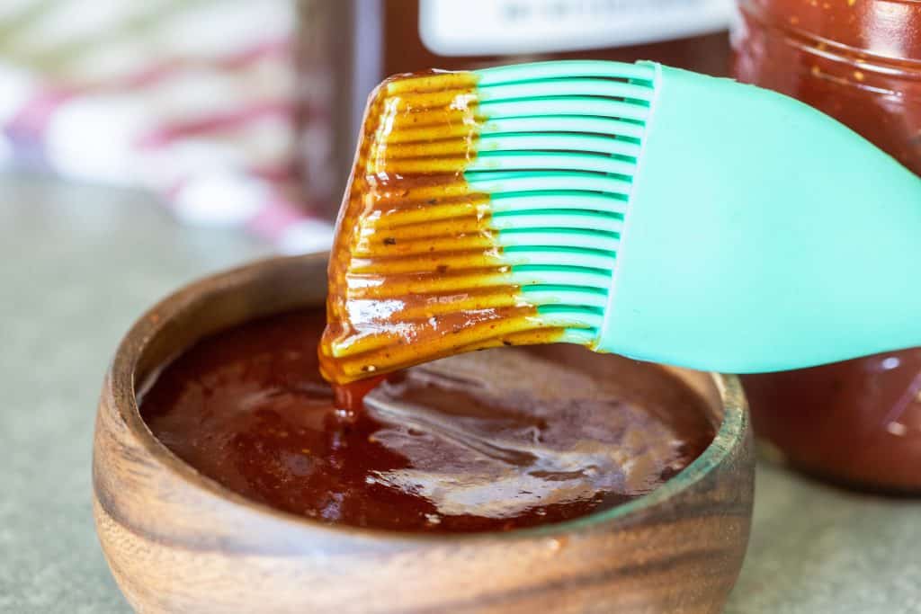 mint colored basting brush dipping in a small wood bowl full of honey barbecue sauce.