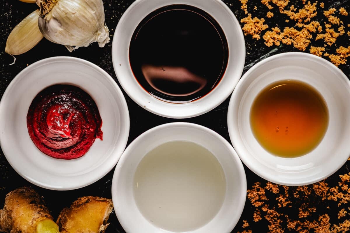 Four white bowls filled with liquids on a countertop next to garlic bulbs and piles of seasonings.