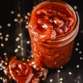 A jar of a thick, red sauce overflowing onto a black countertop sprinkled with salt.
