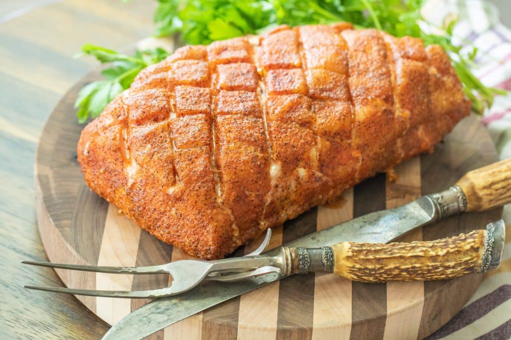 smoked pork loin on a wooden cutting board next to a fork and knife.