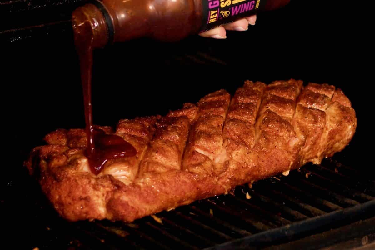 A scored pork roast on the grates of a smoker as a hand pours sauce onto it.