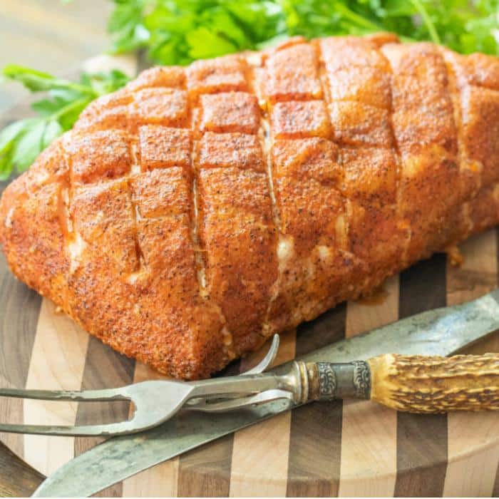 Smoked pork loin on a wooden cutting board next to fresh herbs and a knife and fork.