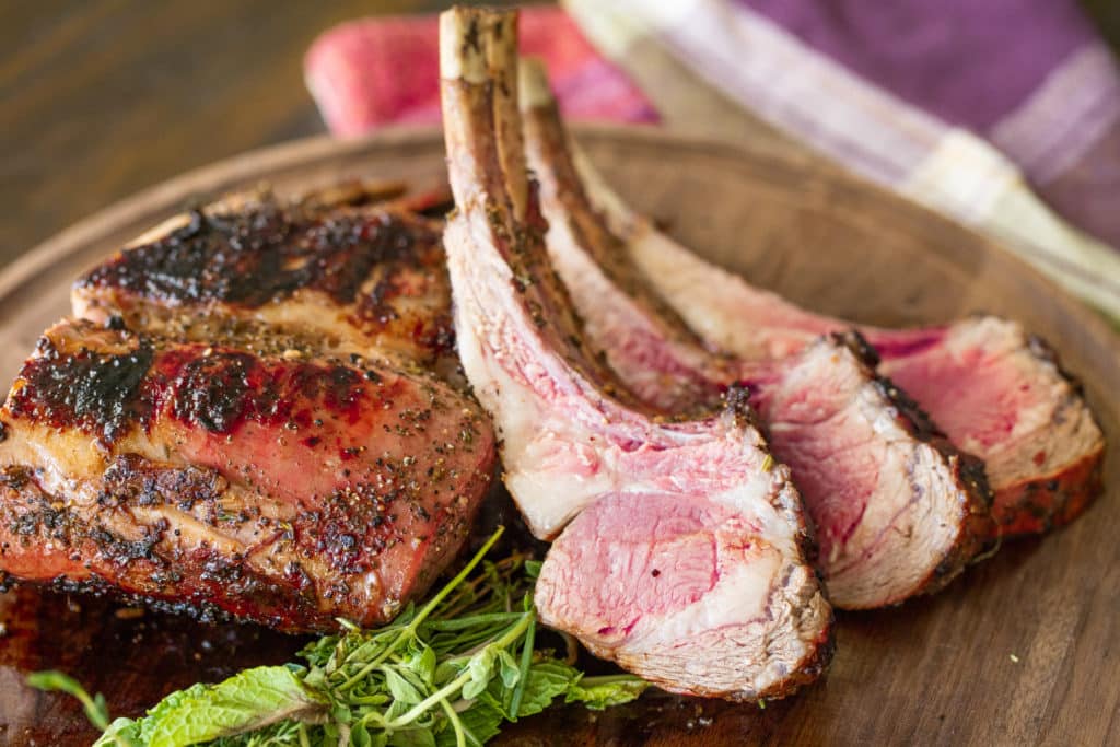 grilled rack of lamb with individual chops arranged on a wood cutting board with herbs.