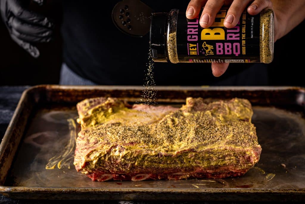 Mustard-slathered beef ribs being sprinkled with Hey Grill Hey Beef Rub.