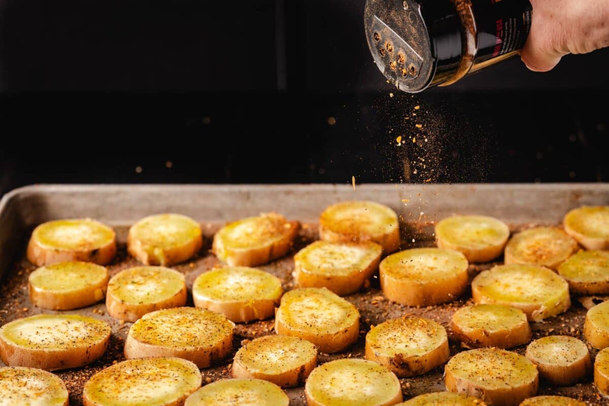 A shaker of seasoning being sprinkled onto sliced veggies on a baking sheet.