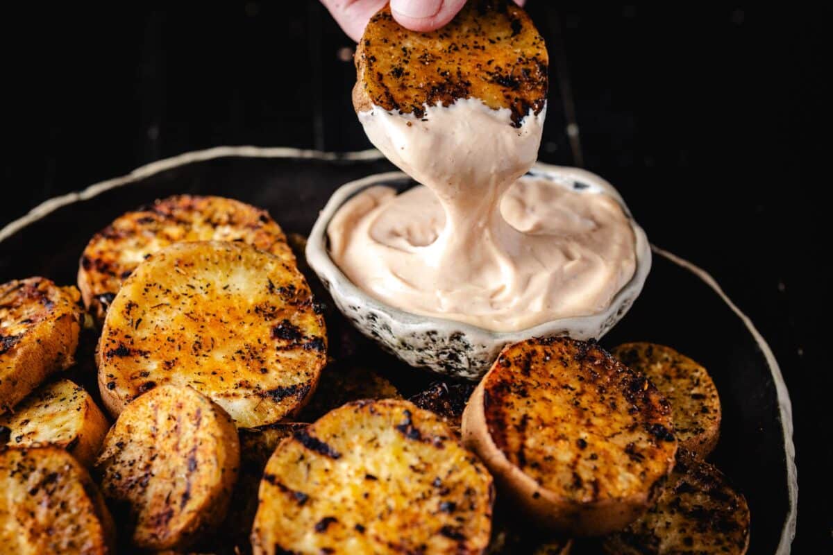 A plate of grilled sweet potatoes and a bowl of dip, with a hand dipping one potato.