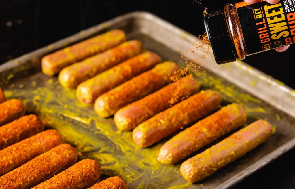 Hot dogs slathered in yellow mustard and being seasoned with Hey Grill Hey Sweet Rub.