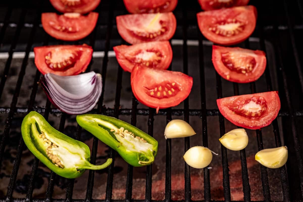 Sliced tomatoes, onions, jalapeños and garlic sitting on the grates of a smoker.