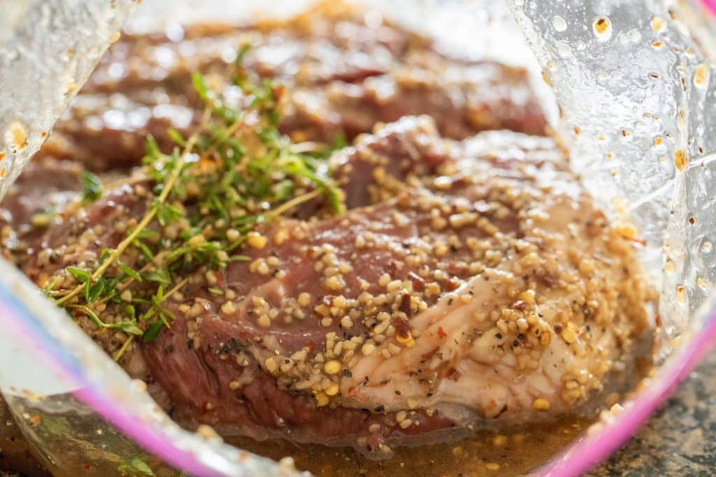 Steak marinating in plastic bag with visible pieces of garlic and thyme.