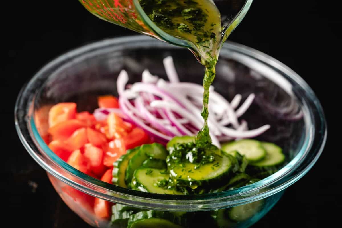 Sliced onions, cucumbers, and tomatoes in a glass bowl with a salad dressing being poured in.