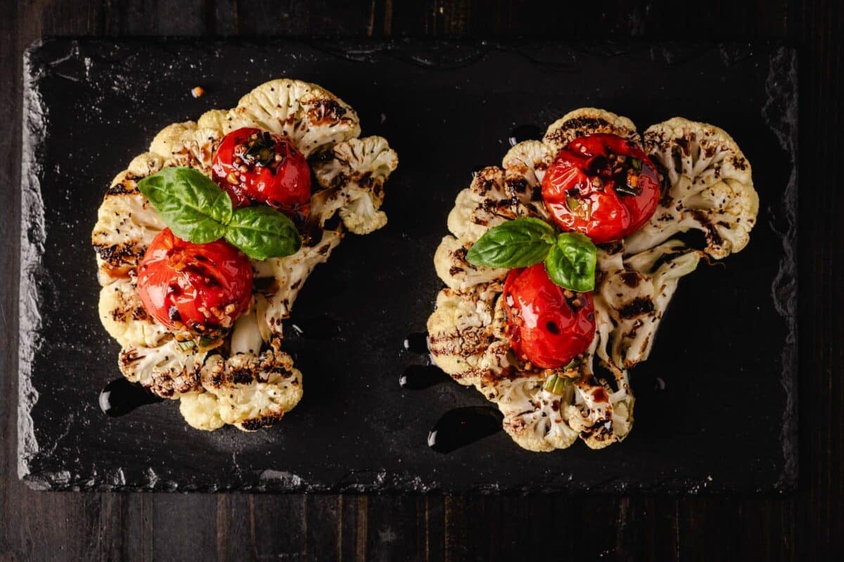 Cauliflower steaks on a black plate topped with tomatoes and basil leaves.