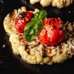 Grilled cauliflower steak topped with tomato and basil on a black serving platter.