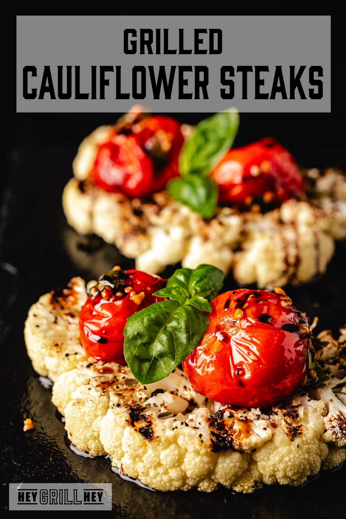 Grilled cauliflower topped with tomato and basil. The text overlay at the top reads "Grilled Cauliflower Steaks," and at the bottom it reads "Hey Grill Hey."