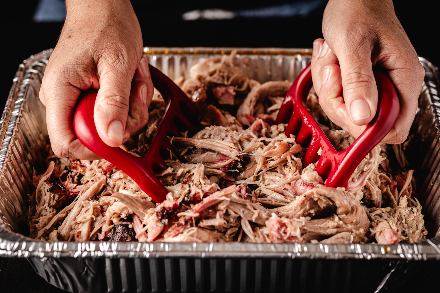Smoked pulled pork being shredded in an aluminum pan.