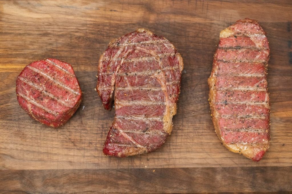Three different cuts of steak grilled and placed on a wooden cutting board.