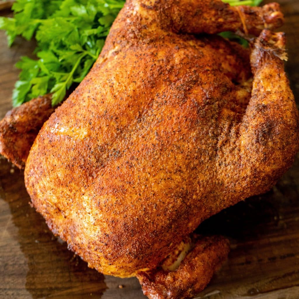 Pollo intero affumicato su un tagliere di legno accanto a erbe fresche.
