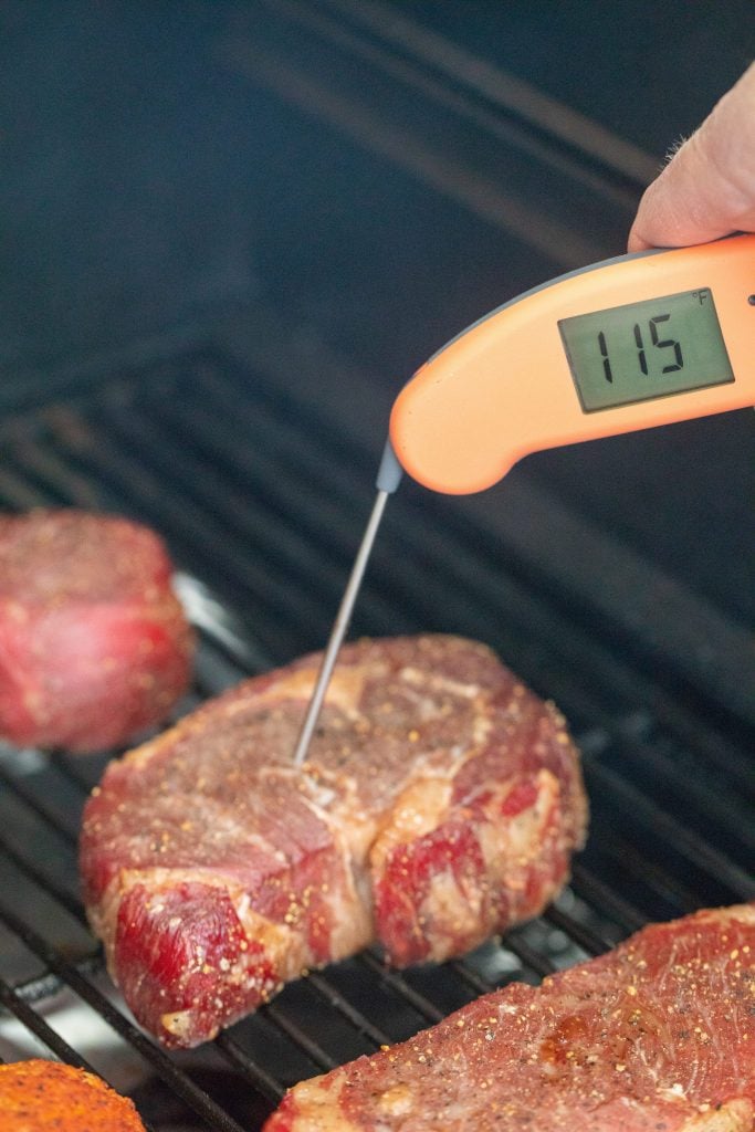 Steaks on the grill grate, as a hand is putting the meat thermometer showing 115 degrees farenheit on the digital thermometer read.