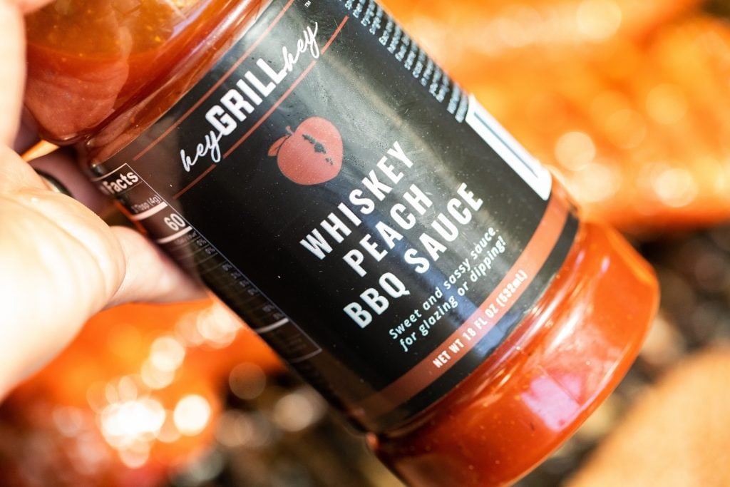 A bottle of Whiskey Peach BBQ sauce being held over smoked pork tenderloins.