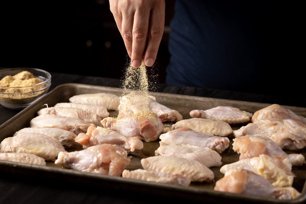 Chicken wings being seasoned with salt, pepper, and garlic powder.