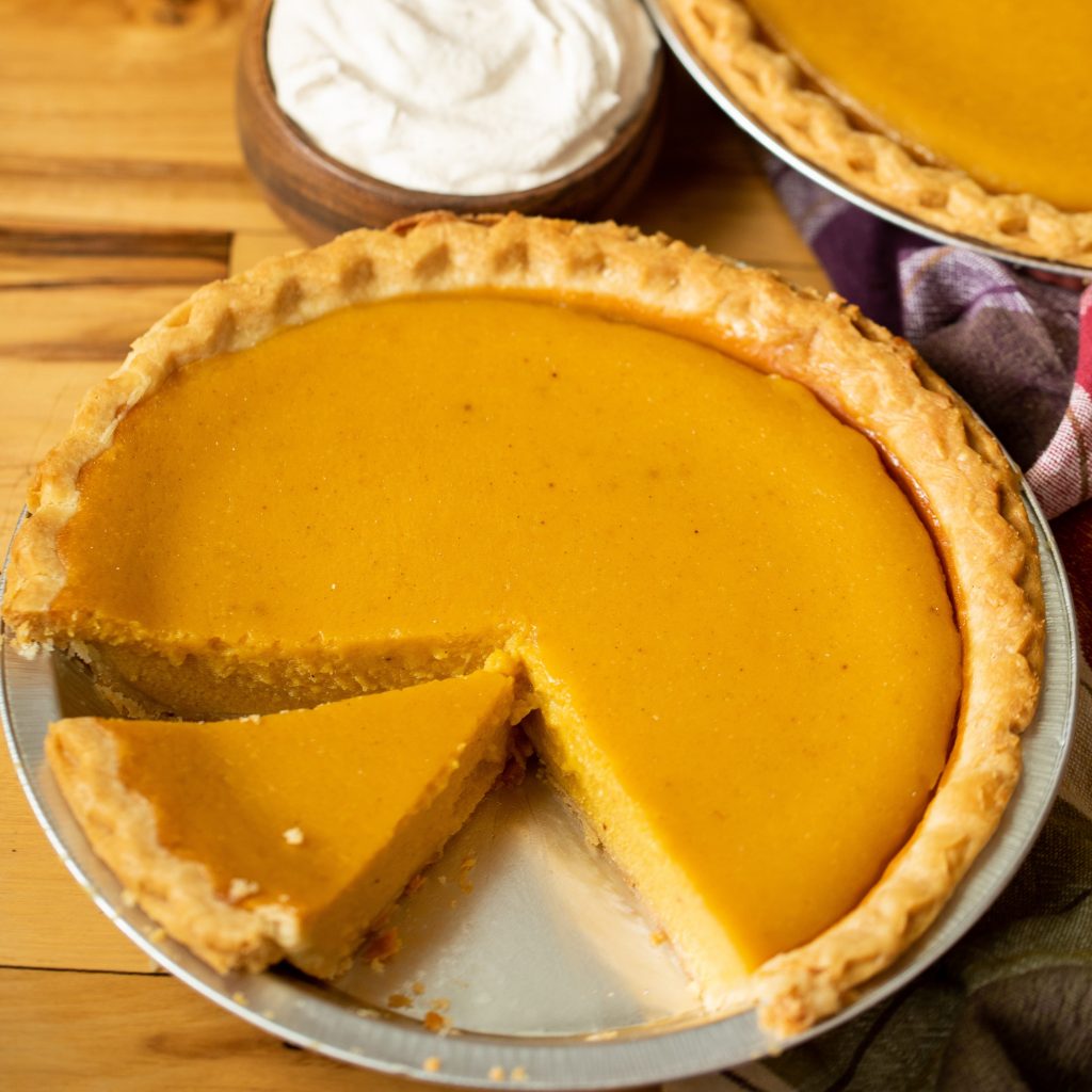 Sliced pumpkin pie in a pie tin next to another whole pie and a small bowl of whipped cream.