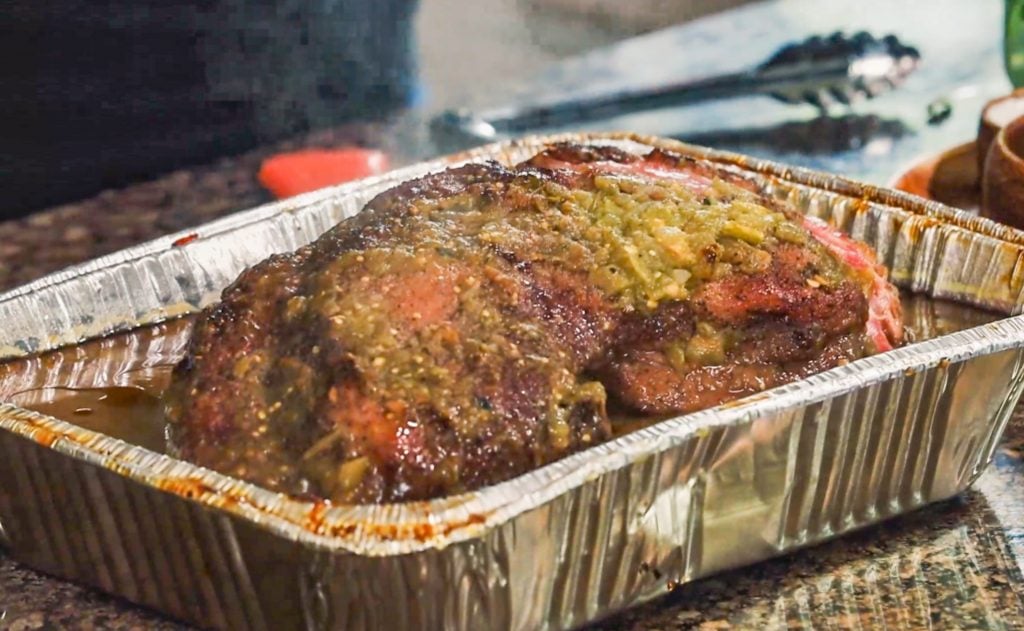 large pork shoulder in disposable aluminum pan resting in sauce and topped with green salsa.