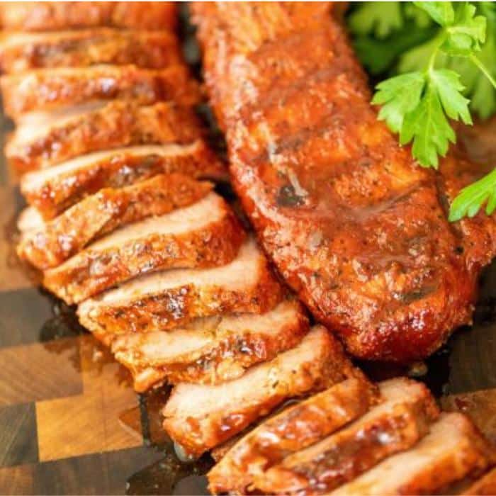 Sliced smoked pork tenderloin next to a whole smoked pork tenderloin on a wooden cutting board.