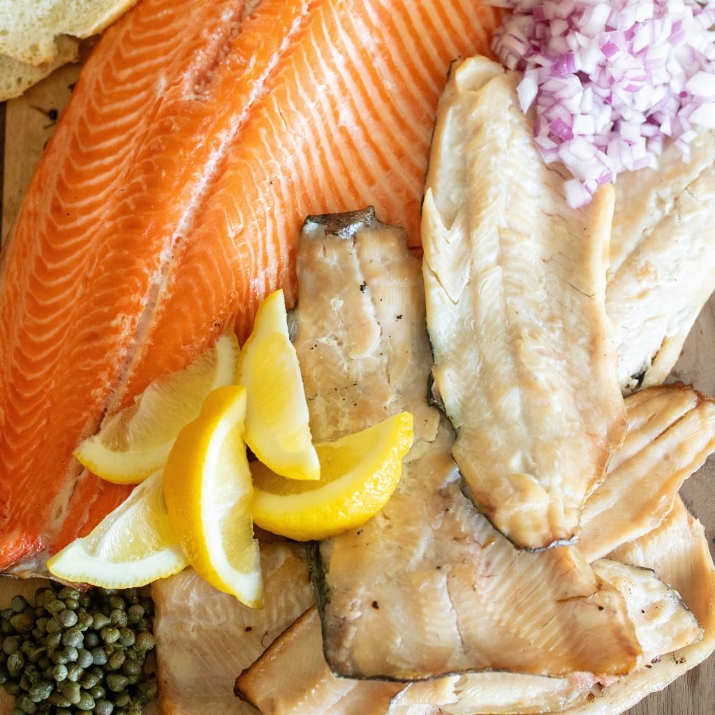 Whole fillets of smoked trout on a wooden board surrounded by sliced baguettes, lemon slices, and chopped onions.