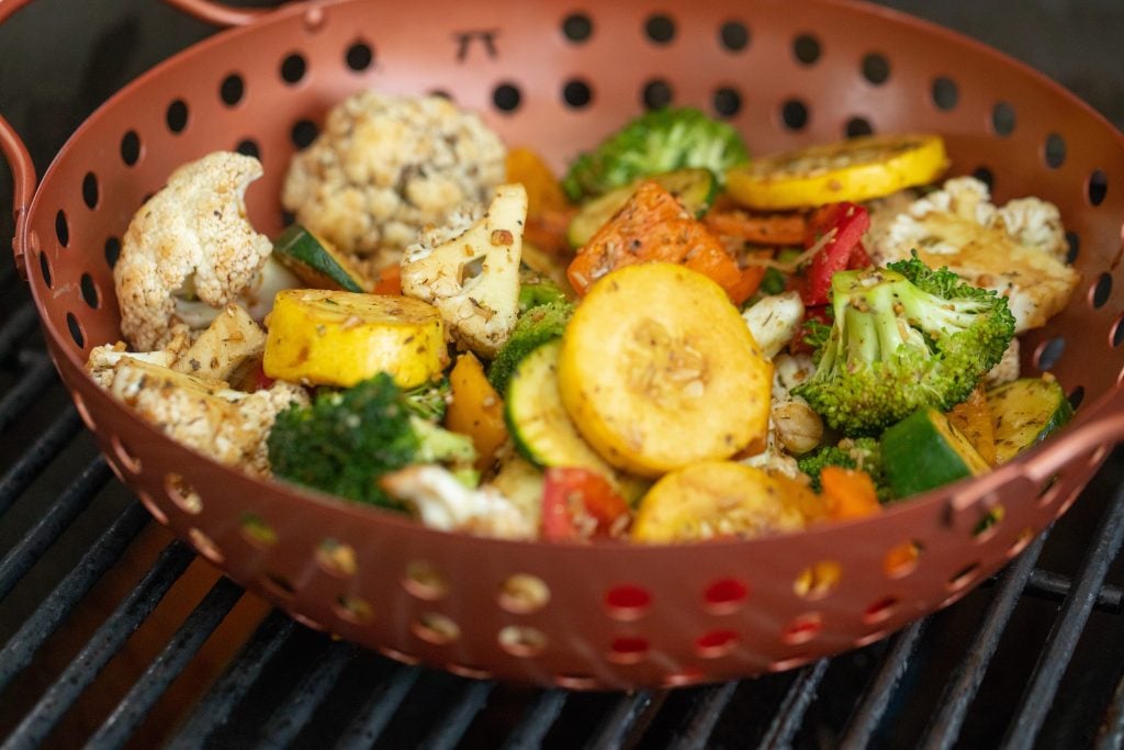 A maroon grill basket bowl with seasoned vegetables in a grill.