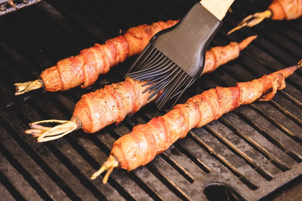 Bacon wrapped carrots on the grill being brushed with maple syrup.
