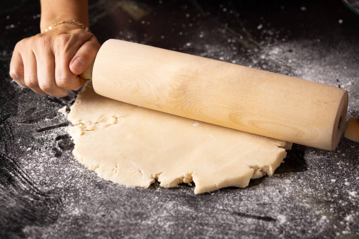 A wooden rolling pin flattening dough on a floured surface.