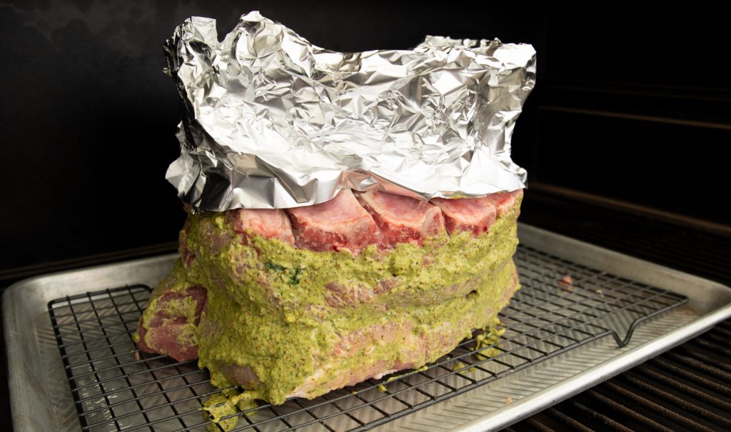 Pork Crown Roast on a rack inside of a smoker with foil covering the bones and an herb paste on the meat.