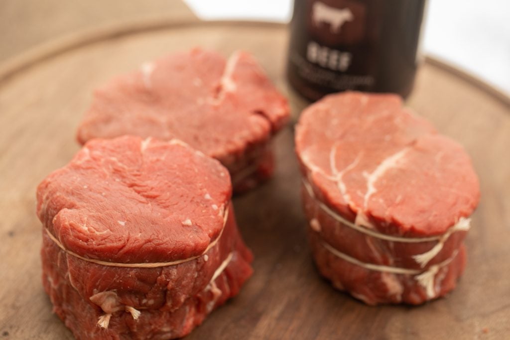 three wrapped filet mignon steaks on a circular wooden cutting board with a bottle of beef rub in the background.