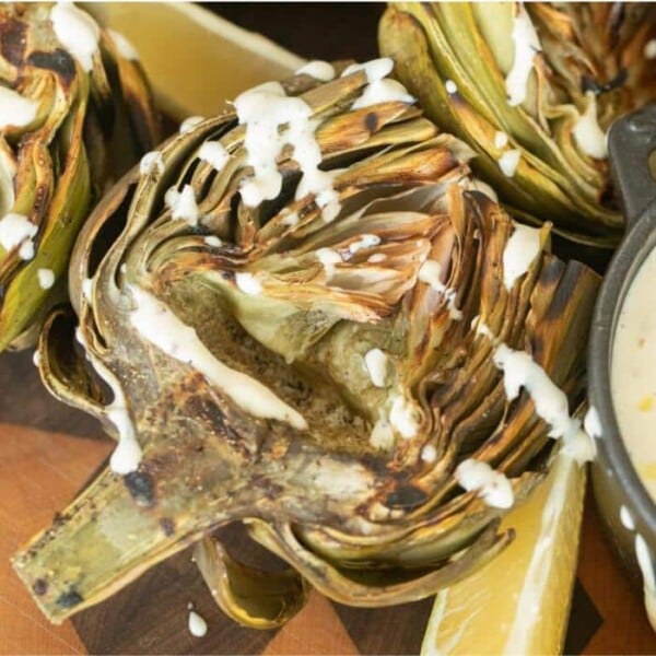 Halved grilled artichokes on a wooden board drizzled with lemon garlic aioli.