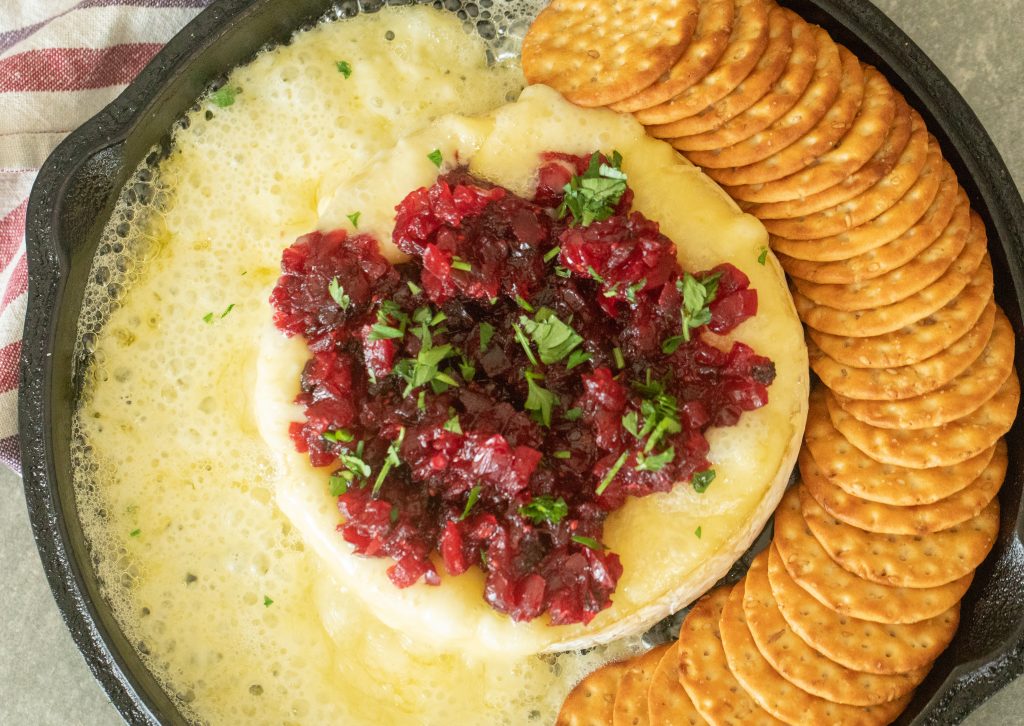 Over head shot of melted grilled brie topped with cranberry jalapeno relish alongside a row of whole grain crackers.