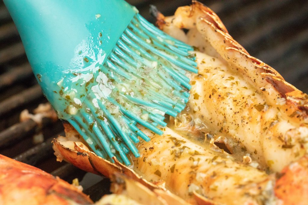 silicon basting brush brushing lemon garlic butter on a lobster tail on a grill.