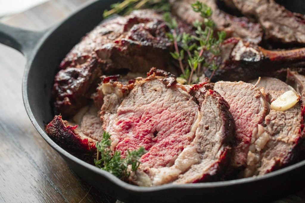 sliced grilled prime rib in a cast iron skillet with fresh herbs and garlic cloves.