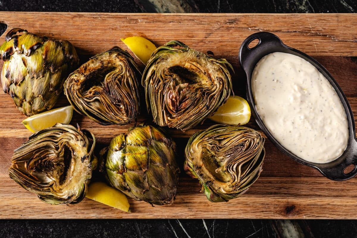 An overhead shot of artichoke halves and a bowl of garlic aioli on a wooden cutting board.