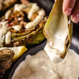 A hand dipping an artichoke leaf into a bowl of dip, in front of a glazed artichoke.