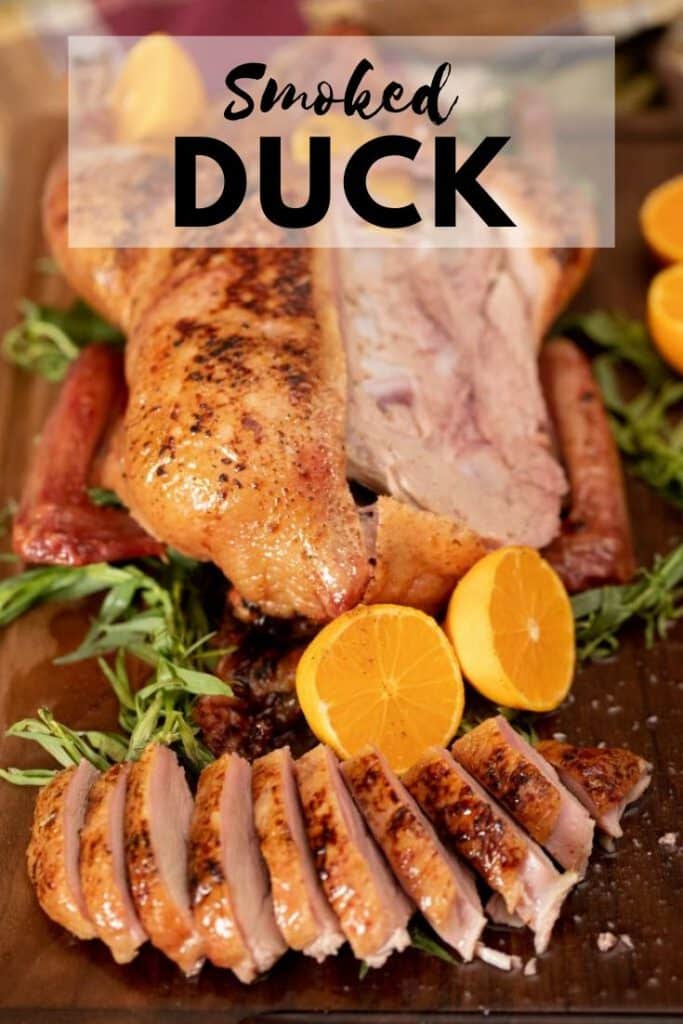 whole smoked sliced duck on a wooden cutting board next to fresh herbs and halved oranges. Text overlay reads "Smoked Duck."