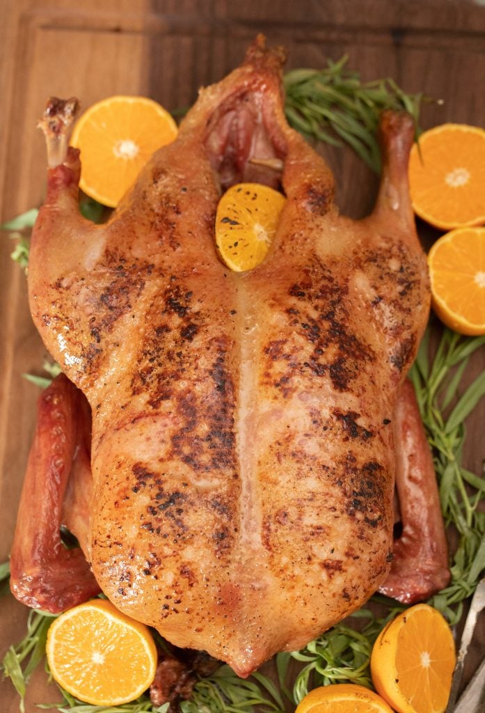 smoked whole duck on a wooden cutting board with fresh herbs and sliced oranges.