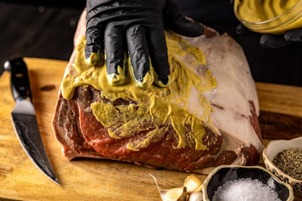 New york strip roast being rubbed with yellow mustard.