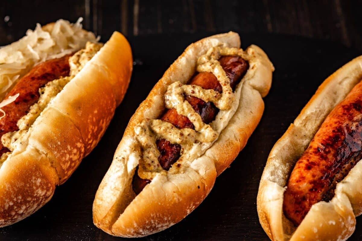 A grilled bratwurst on a rolled drizzled with spicy mustard, another topped with sauerkraut, and one plain.