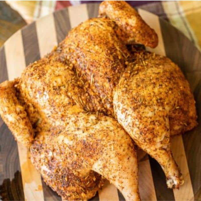 whole cooked chicken, covered with seasoning and resting on a wooden cutting board. 