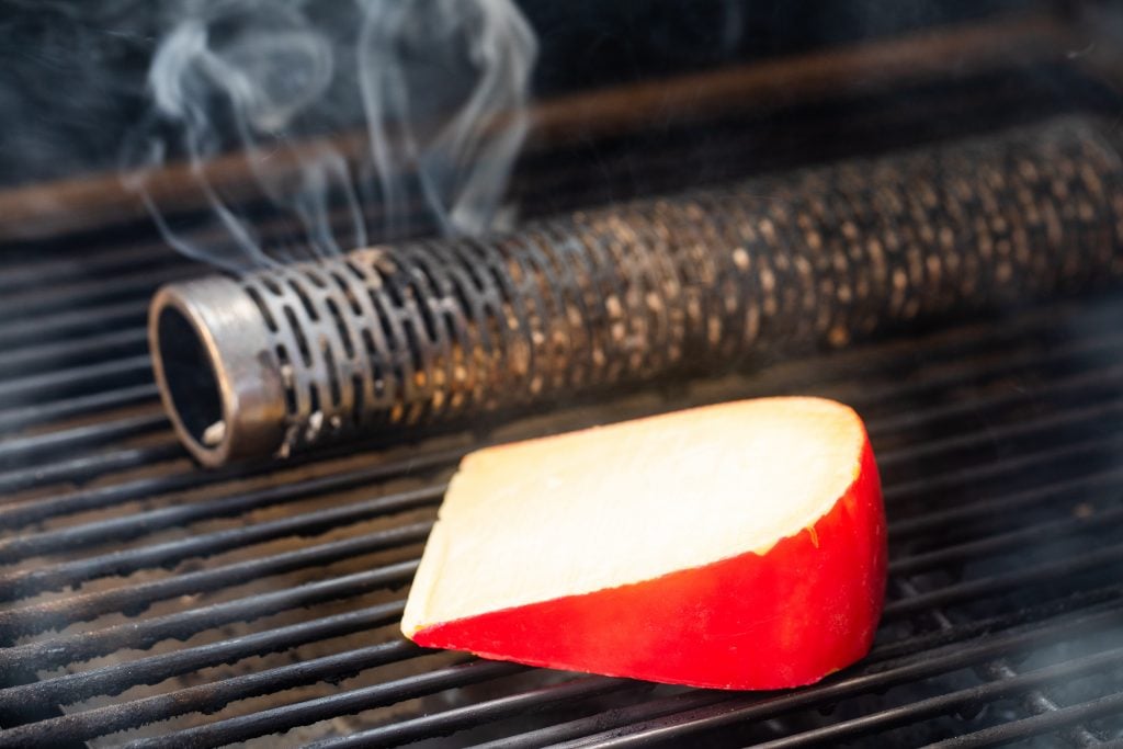 gouda cheese on the grill grates of a smoker next to a smoke tube.